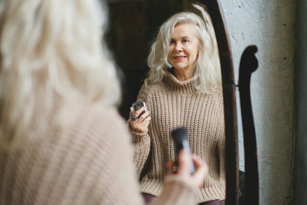 pexels-photo-5682592-5682592 A senior woman in a cozy sweater smiles while applying makeup in front of a mirror.